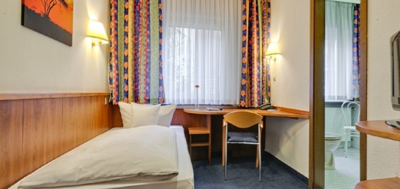 Ein gemütliches Einzelzimmer mit einem Bett, Schreibtisch und Stuhl. Bunte Vorhänge und ein Bild schmücken den Raum. Ein Badezimmer ist im Hintergrund sichtbar., © ARNOTEL GmbH