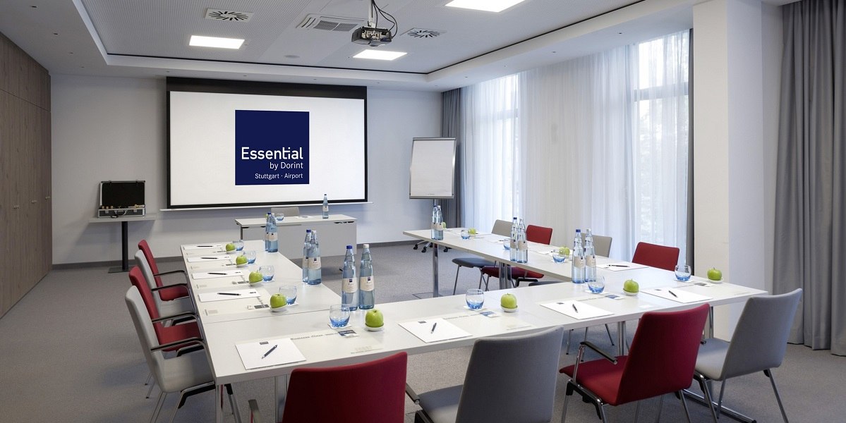 Meeting room with U-shaped table arrangement, chairs, water bottles, apples and writing materials. Projector shows 'Essential by Dorint Stuttgart Airport'., &copy; Essential by Dorint