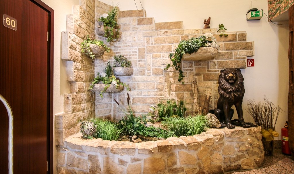 Steinwand mit Pflanzenarrangement und Löwenstatue in einer Halle. Tür mit Nummer 60 und Notausgangsschild sichtbar., © Ulli Kellenbenz