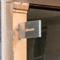 Ein Metallschild mit der Aufschrift 'abnorm' h&auml;ngt an einer Wand. Daneben ist ein Kunstplakat zu sehen., &copy; SMG, Sarah Schmid