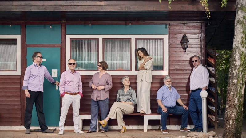 Sieben Personen posieren lässig vor einem Holzgebäude mit türkisfarbener Tür. Einige stehen, andere sitzen auf Bänken., © Martin Sigmund Sieben Personen posieren lässig vor einem Holzgebäude mit türkisfarbener Tür. Einige stehen, andere sitzen auf Bänken., © Martin Sigmund