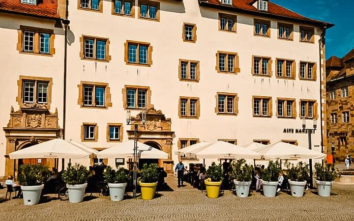Ein historisches Gebäude mit roten Dächern und vielen Fenstern. Vor dem Gebäude stehen Sonnenschirme und Pflanzenkübel auf einem gepflasterten Platz., © Alte Kanzlei Ein historisches Gebäude mit roten Dächern und vielen Fenstern. Vor dem Gebäude stehen Sonnenschirme und Pflanzenkübel auf einem gepflasterten Platz., © Alte Kanzlei