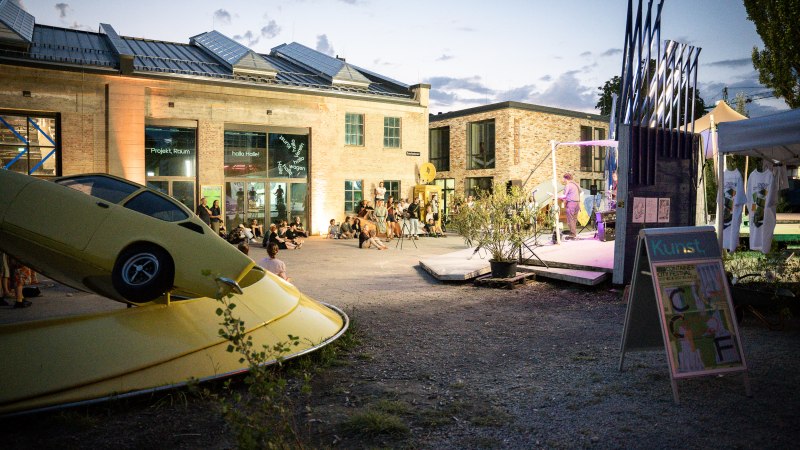 Abendveranstaltung im Kunstverein Wagenhalle mit Kunstinstallation eines gelben Autos und Menschen, die auf dem Platz sitzen und einer Darbietung zuschauen., © Atelier Oradoro