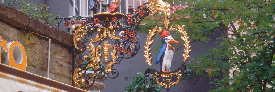 An artistically decorated sign with a penguin and figures, surrounded by floral ornaments, hangs on a wall on Eugensplatz., &copy; Stuttgart-Marketing GmbH