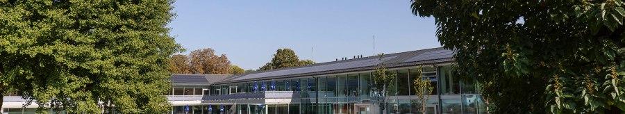 The picture shows the outdoor area of the Mineralbad Berg with a pool, surrounded by modern buildings and green trees under a clear sky., © Stuttgarter Bäder