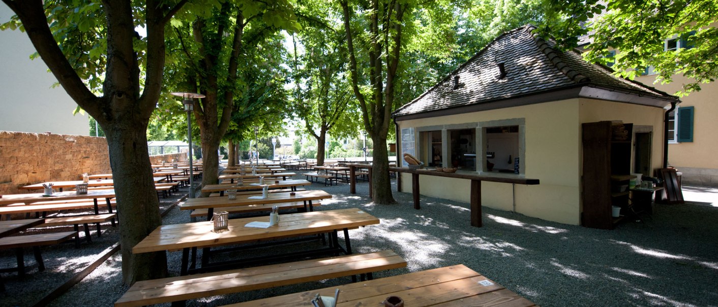 Ein ruhiger Biergarten mit Holztischen und Bänken unter Bäumen, neben einem kleinen Gebäude. Die Sonne scheint durch das Blätterdach., © Ruthenberg&Schlatter GbR Ein ruhiger Biergarten mit Holztischen und Bänken unter Bäumen, neben einem kleinen Gebäude. Die Sonne scheint durch das Blätterdach., © Ruthenberg&Schlatter GbR