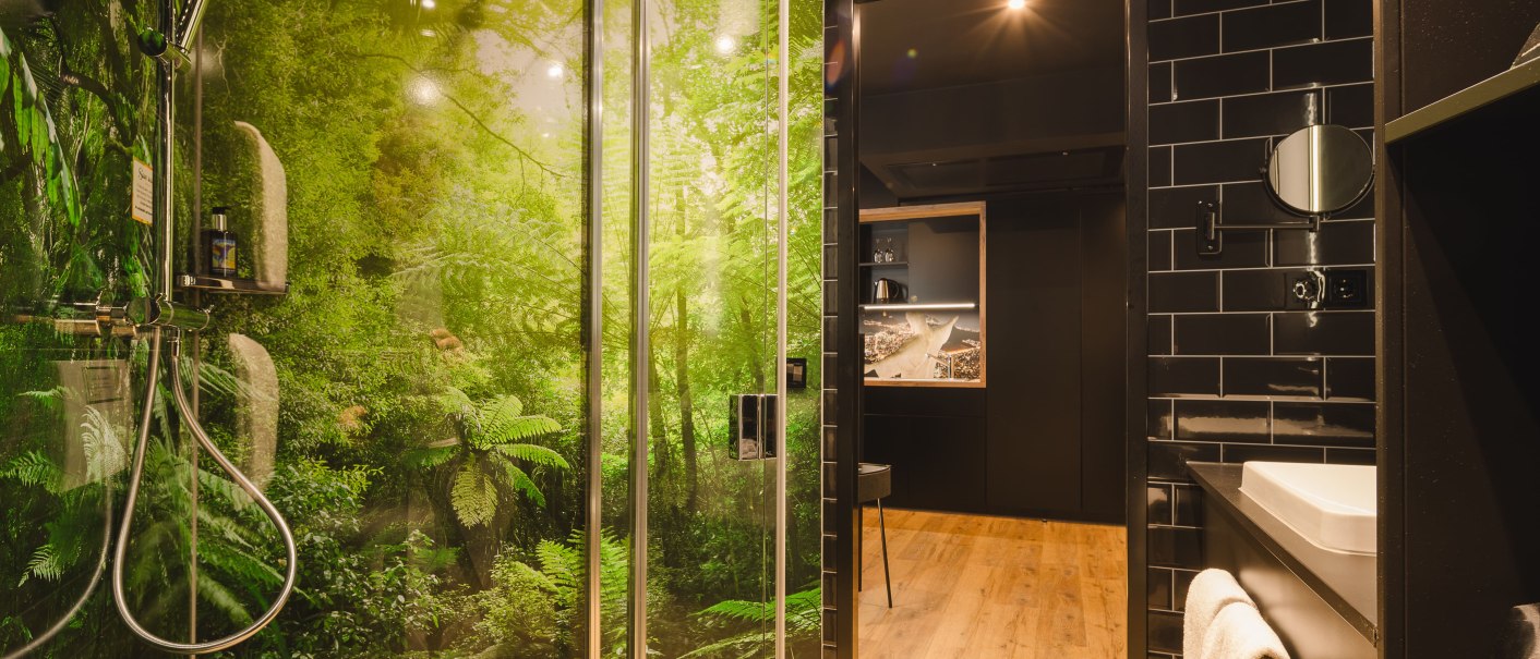 Modernes Badezimmer mit Dschungelmotiv in der Dusche, schwarzen Fliesen und Holzfußboden. Ein Waschbecken mit Handtuch ist sichtbar., © TOMAS