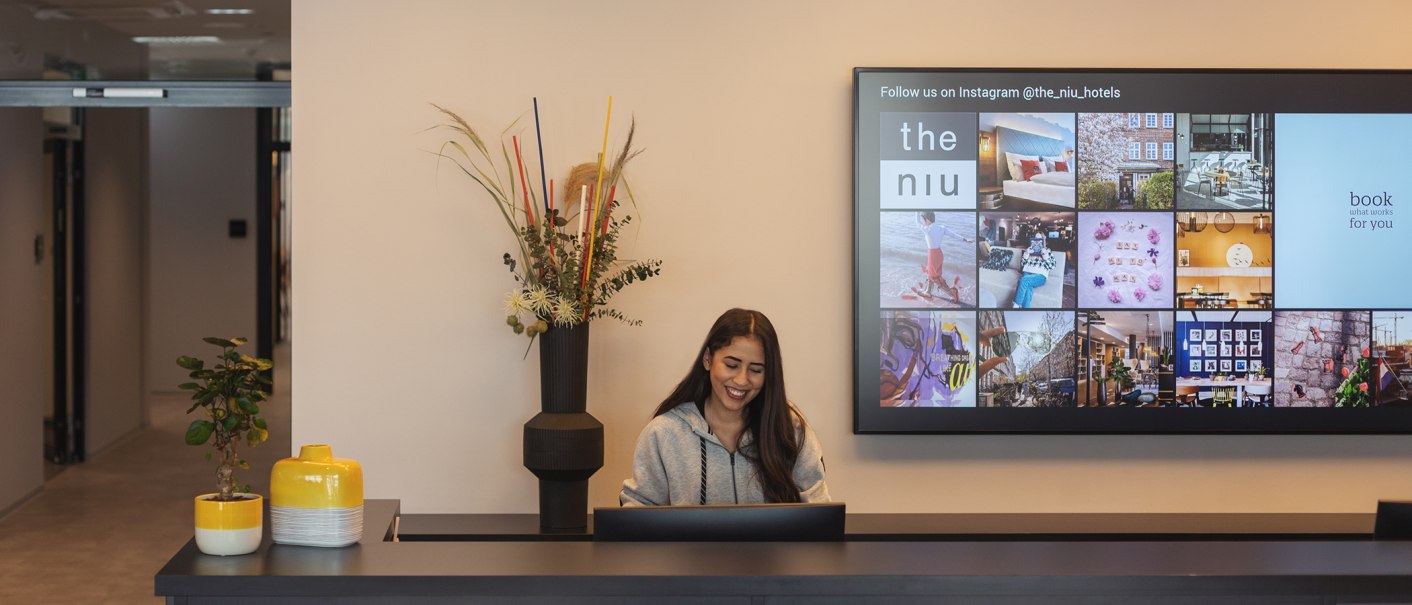 A woman at a hotel front desk, smiling, with plants and a screen displaying Instagram photos in the background., © the niu Form - Novum Hospitality A woman at a hotel front desk, smiling, with plants and a screen displaying Instagram photos in the background., © the niu Form - Novum Hospitality