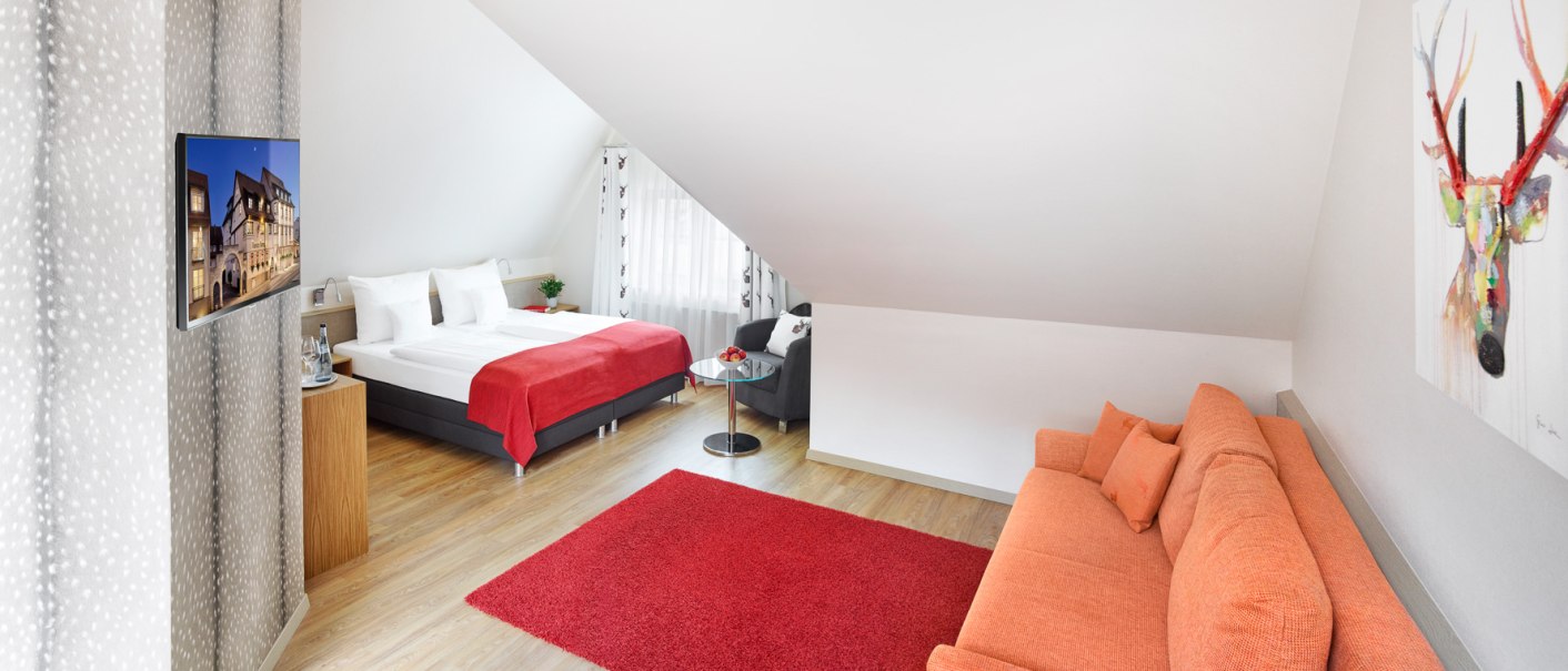 Helle Junior Suite mit Doppelbett, rotem Teppich, oranger Couch und Hirschbild an der Wand. Ein Fernseher und ein kleiner Tisch sind ebenfalls vorhanden., © Hirsch Hotel