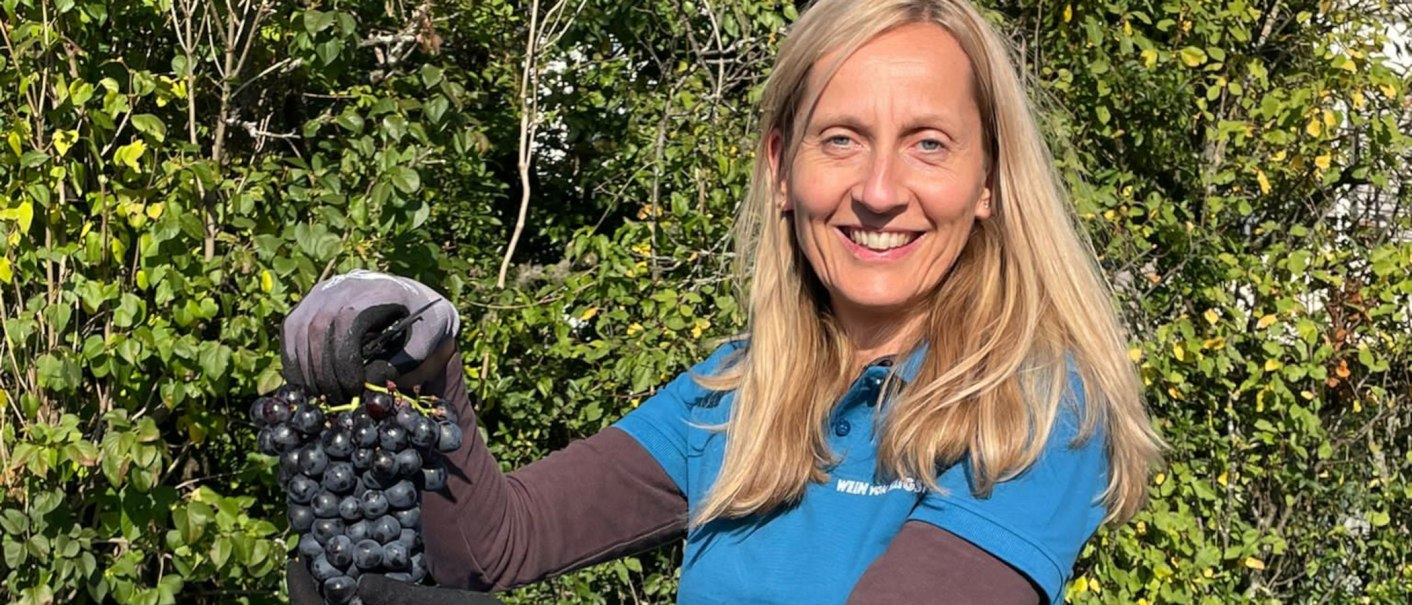Frau mit blonden Haaren h&auml;lt Trauben in der Hand, l&auml;chelt in die Kamera. Sie tr&auml;gt ein blaues Shirt, im Hintergrund sind gr&uuml;ne B&uuml;sche zu sehen., &copy; weinvomhaigst