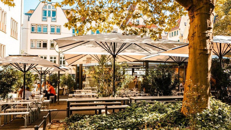 Sonniger Café-Außenbereich mit weißen Sonnenschirmen, Holztischen und Pflanzen. Menschen sitzen entspannt unter den Schirmen. Gebäude im Hintergrund., © Stuttgart Marketing GmbH, Sarah Schmid Sonniger Café-Außenbereich mit weißen Sonnenschirmen, Holztischen und Pflanzen. Menschen sitzen entspannt unter den Schirmen. Gebäude im Hintergrund., © Stuttgart Marketing GmbH, Sarah Schmid