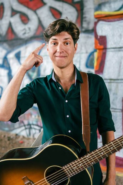 A man with a guitar stands in front of a graffiti wall and points to his head with his finger. He is wearing a dark green shirt and smiling slightly., &copy; Rosenau Kultur e.V.