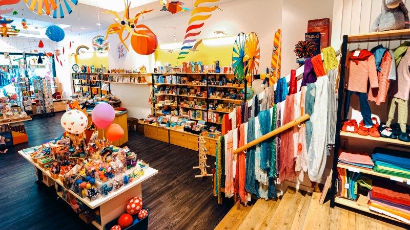 Ein bunter Spielzeugladen mit Regalen voller Spielsachen, Kleidung und Dekorationen. Bunte Schals und Spielzeuge sind prominent ausgestellt., &copy; 1000sch&ouml;n