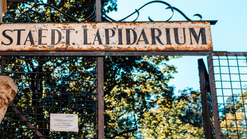 Eingang zum St&auml;dtischen Lapidarium mit einem alten, verwitterten Schild und einer Steinskulptur. Im Hintergrund sind B&auml;ume zu sehen., &copy; Stuttgart Marketing GmbH, Sarah Schmid