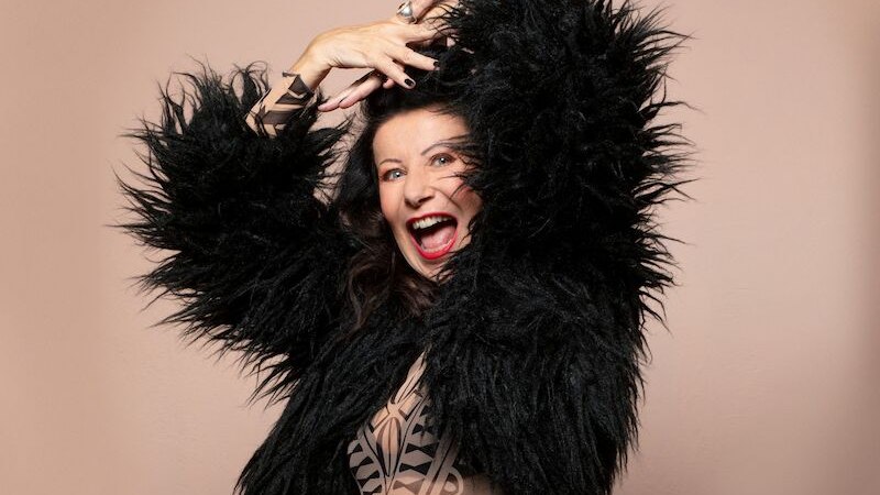 Eine Frau in einem schwarzen, flauschigen Outfit posiert fröhlich vor einem beigen Hintergrund. Sie lacht und hebt die Arme in einer dynamischen Pose., © Theaterhaus Stuttgart e.V. Eine Frau in einem schwarzen, flauschigen Outfit posiert fröhlich vor einem beigen Hintergrund. Sie lacht und hebt die Arme in einer dynamischen Pose., © Theaterhaus Stuttgart e.V.