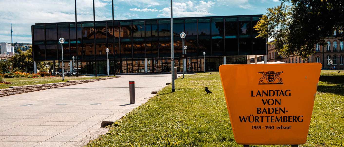 The state parliament of Baden-Württemberg with a modern glass façade and a yellow sign in the foreground. The television tower can be seen in the background., © Stuttgart-Marketing GmbH, Sarah Schmid