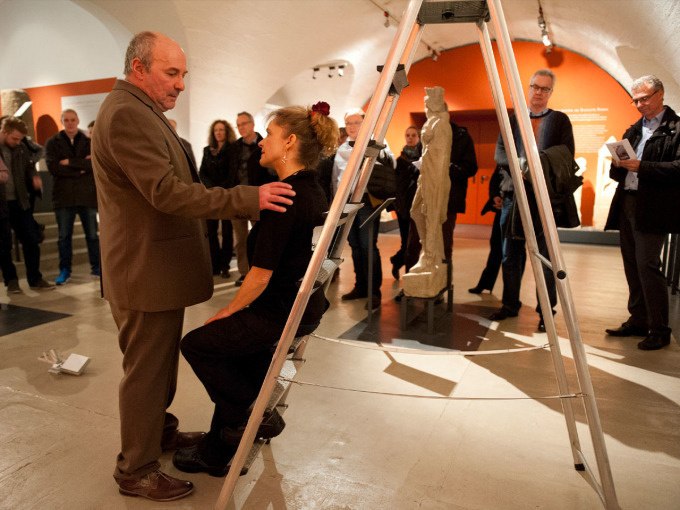 A woman sits on a ladder in a museum, surrounded by spectators. A man stands in front of her, while statues can be seen in the background., © Landesmuseum Württemberg A woman sits on a ladder in a museum, surrounded by spectators. A man stands in front of her, while statues can be seen in the background., © Landesmuseum Württemberg