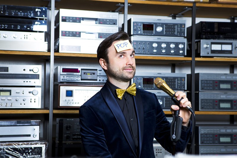A man in a suit with a golden bow tie is holding a microphone. A note with 'POP' is stuck to his forehead. Many audio devices can be seen in the background., &copy; Theaterhaus Stuttgart e.V.