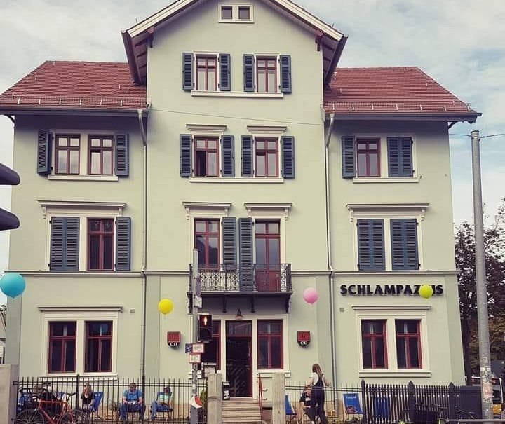 Three-storey building with 'Schlampazius' lettering, colorful balloons and people in front of the entrance. Historic architecture with red window frames., &copy; Mora F&uuml;tterer
