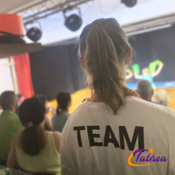 Person mit TEAM-Shirt steht vor einer B&uuml;hne, auf der eine Auff&uuml;hrung stattfindet. Zuschauer sitzen im Hintergrund. Logo 'Tathea' unten rechts., &copy; Tathea - Lena Oschowitzer