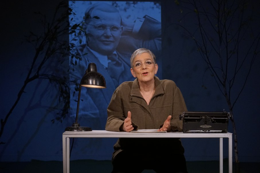 Eine Frau sitzt an einem Tisch mit einer Schreibmaschine und einer Lampe. Im Hintergrund ist ein projiziertes Bild eines Mannes zu sehen., © Katja Ritter Eine Frau sitzt an einem Tisch mit einer Schreibmaschine und einer Lampe. Im Hintergrund ist ein projiziertes Bild eines Mannes zu sehen., © Katja Ritter