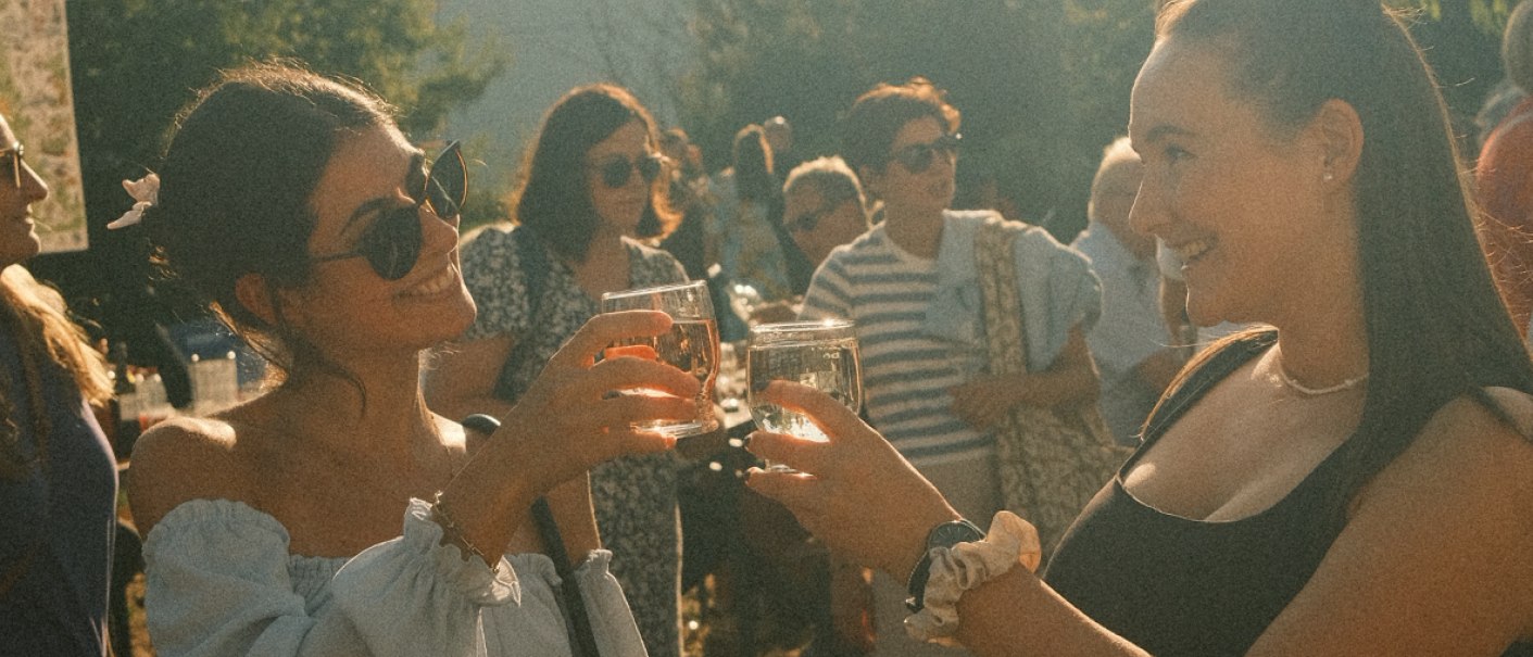 Zwei Frauen stoßen mit Weingläsern an, während sie in einer fröhlichen Menschenmenge im Freien stehen., © MyDegerloch Zwei Frauen stoßen mit Weingläsern an, während sie in einer fröhlichen Menschenmenge im Freien stehen., © MyDegerloch