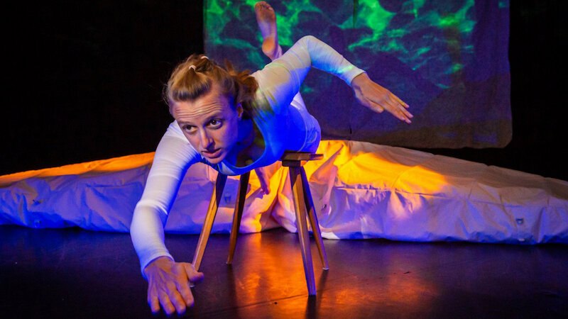 Eine Frau in wei&szlig;em Outfit balanciert auf einem Hocker, umgeben von bunten Lichteffekten, die eine Unterwasserwelt darstellen., &copy; FITZ Das Theater animierter Formen