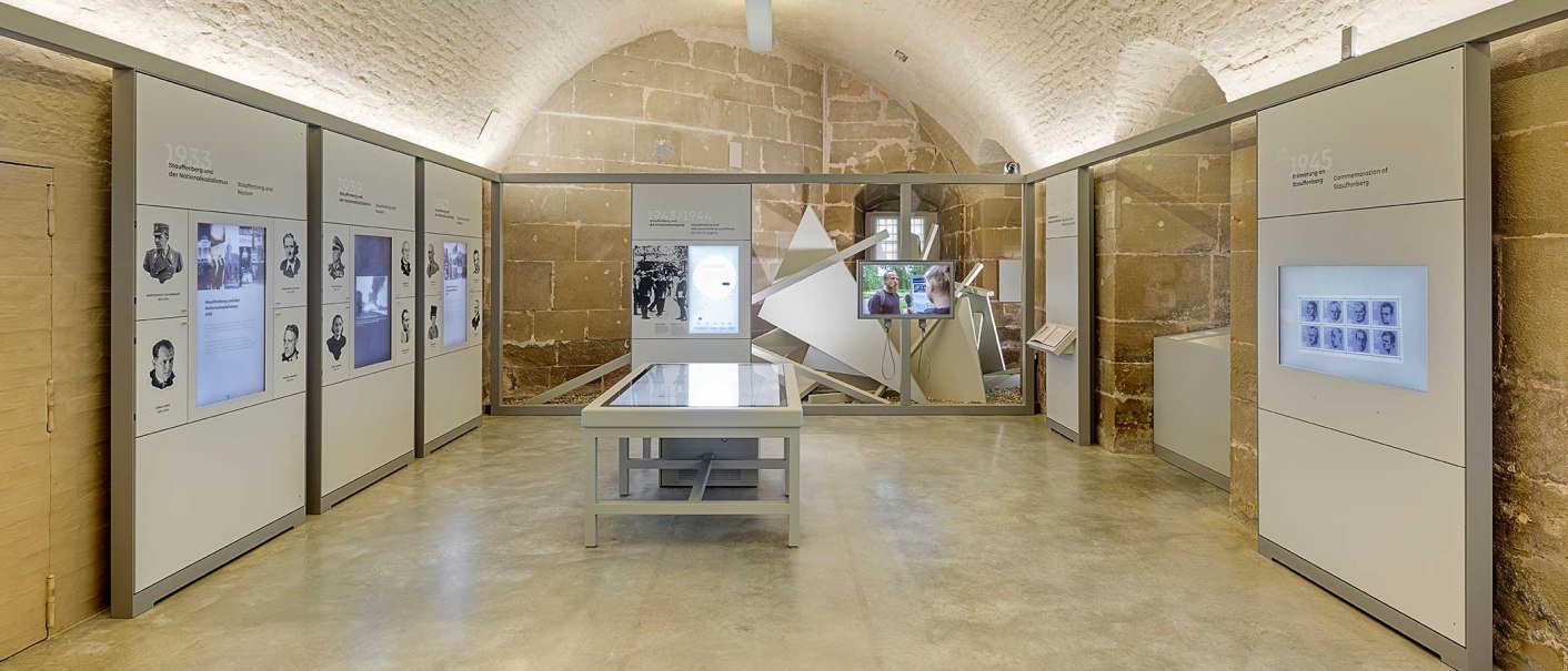 Innenansicht der Stauffenberg-Erinnerungsstätte mit modernen Ausstellungswänden, interaktiven Displays und einem zentralen Tisch in einem Gewölberaum., © Haus der Geschichte Baden-Württemberg