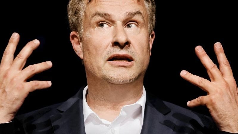 Ein Mann in einem Anzug gestikuliert mit erhobenen Händen vor einem schwarzen Hintergrund., © Stuttgarter Kammerorchester e.V.