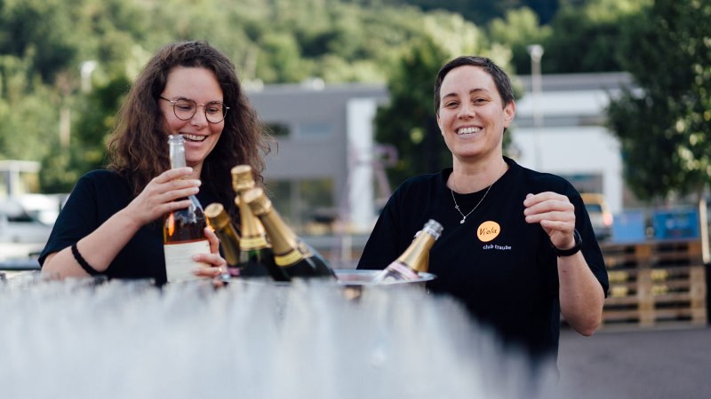 Two people are laughing and holding bottles of wine. They are standing outside, surrounded by trees and buildings in the background., © Bernhard Kahrmann Two people are laughing and holding bottles of wine. They are standing outside, surrounded by trees and buildings in the background., © Bernhard Kahrmann