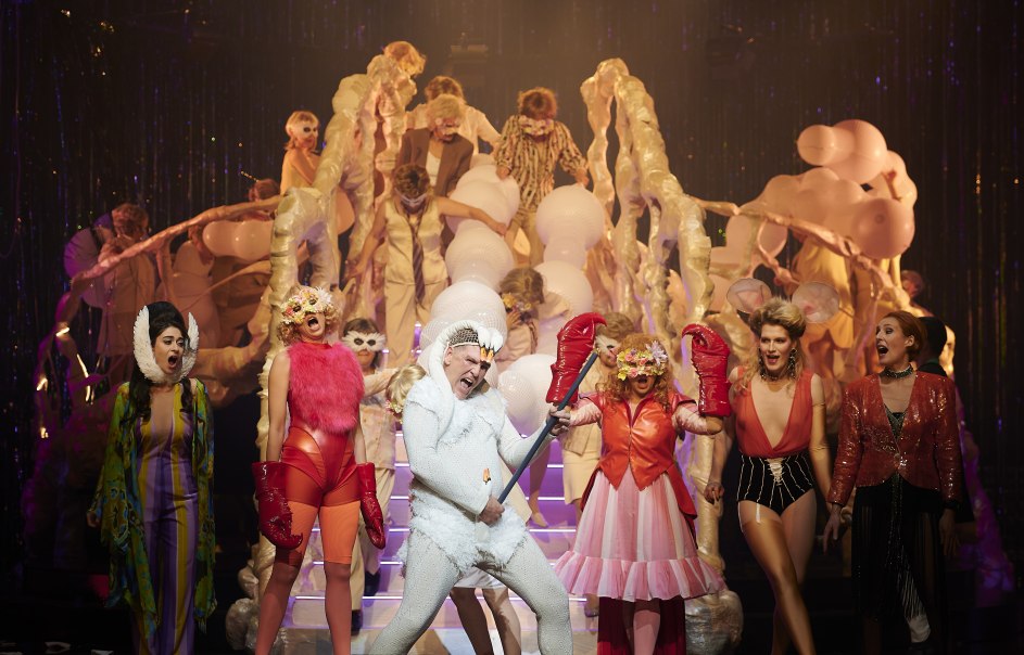 Bunt kost&uuml;mierte Darsteller auf einer B&uuml;hne, umgeben von einer skulpturalen Kulisse. Die Szene wirkt lebhaft und theatralisch., &copy; Matthias Baus