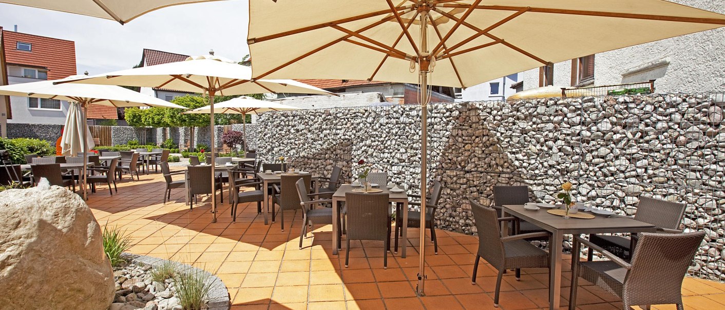 Sonnige Terrasse mit Tischen und Stühlen unter großen Sonnenschirmen, umgeben von einer Steinmauer. Gepflasterter Boden und dekorative Pflanzen., © Hotel Hirsch