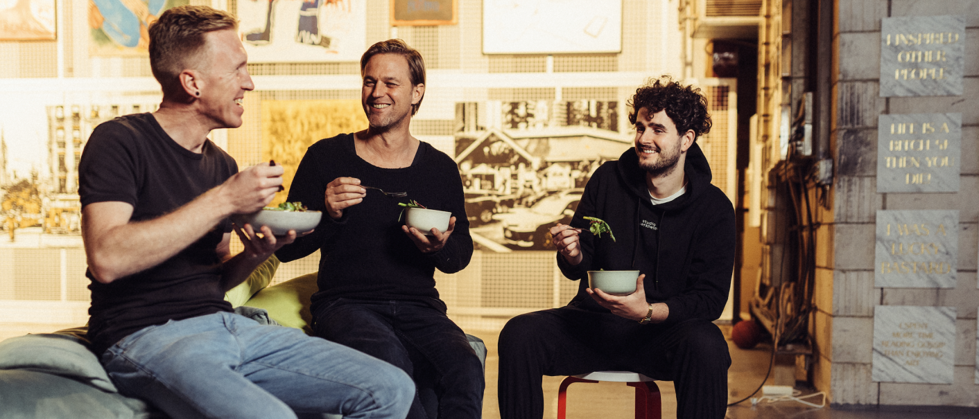 Three men sit together laughing and eating salad in a cozy, artistically designed room., © Vhy! GmbH