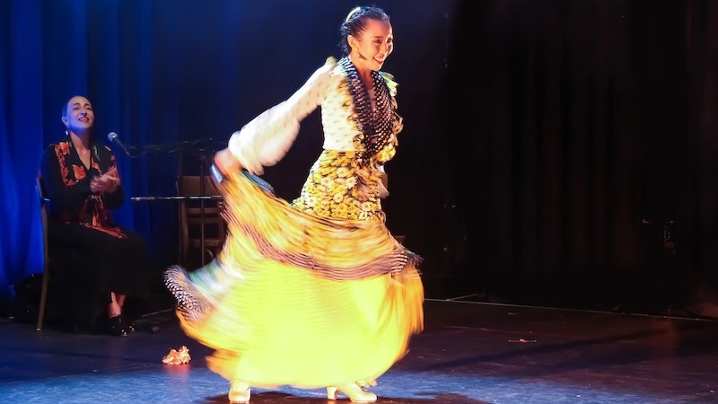 A flamenco dancer in a bright yellow skirt dances on stage while a musician applauds in the background., &copy; Stuttgarter Flamenco Festival gUG