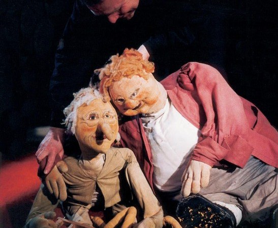 Ein Puppenspieler manipuliert zwei große Puppen, die auf dem Boden sitzen. Eine Puppe trägt eine rote Jacke, die andere eine beige Kleidung., © FITZ Das Theater animierter Formen Ein Puppenspieler manipuliert zwei große Puppen, die auf dem Boden sitzen. Eine Puppe trägt eine rote Jacke, die andere eine beige Kleidung., © FITZ Das Theater animierter Formen