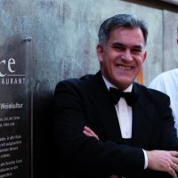 Two men stand in front of a sign for the Délice restaurant. One is wearing a suit, the other a chef's jacket., © Delice Two men stand in front of a sign for the Délice restaurant. One is wearing a suit, the other a chef's jacket., © Delice
