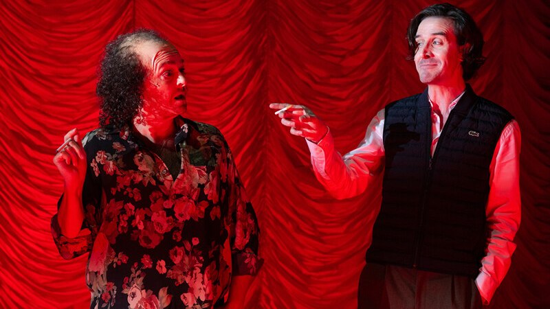 Zwei Männer stehen vor einem roten Vorhang. Der linke trägt ein Blumenhemd, der rechte eine Weste. Beide gestikulieren lebhaft., © Württembergische Staatstheater Stuttgart