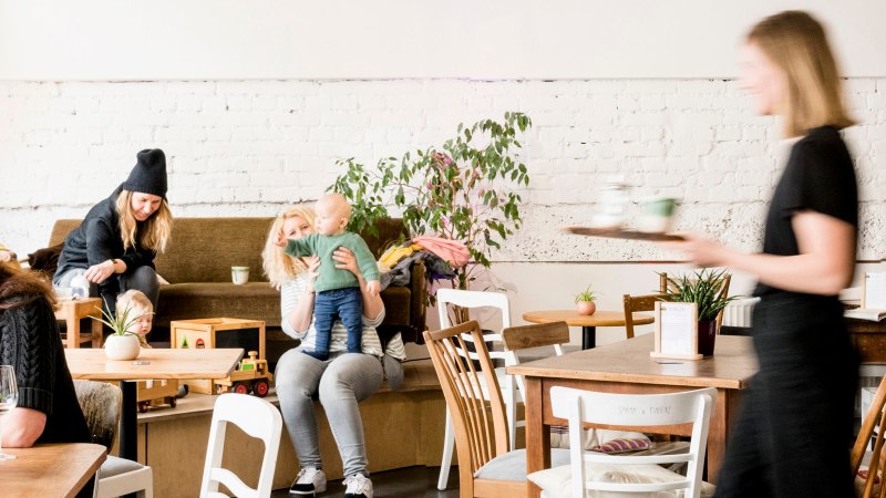Gemütliches Café mit spielenden Kindern und Erwachsenen. Eine Bedienung serviert Getränke. Pflanzen und Holzmöbel schaffen eine einladende Atmosphäre., © Sven Weber