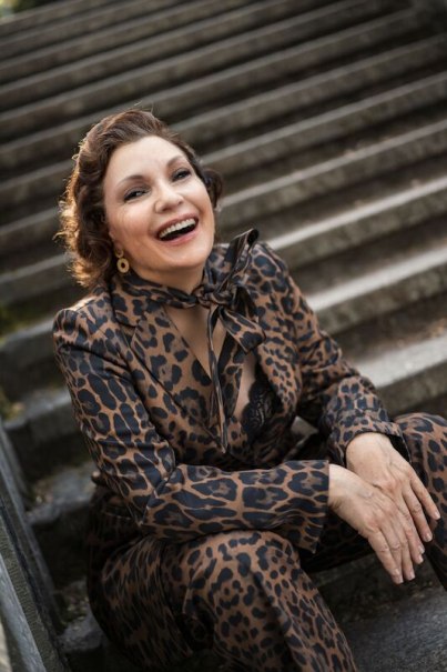 Eine Frau in einem Leopardenmuster-Anzug sitzt l&auml;chelnd auf einer Steintreppe und strahlt Freude aus., &copy; Theaterhaus Stuttgart e.V.