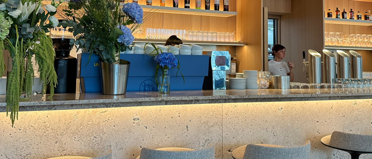 Moderne Bar mit blauen und weißen Blumenarrangements, Getränken im Regal und grauen Barhockern. Eine Person steht hinter der Theke., © Stuttgart-Marketing GmbH Moderne Bar mit blauen und weißen Blumenarrangements, Getränken im Regal und grauen Barhockern. Eine Person steht hinter der Theke., © Stuttgart-Marketing GmbH
