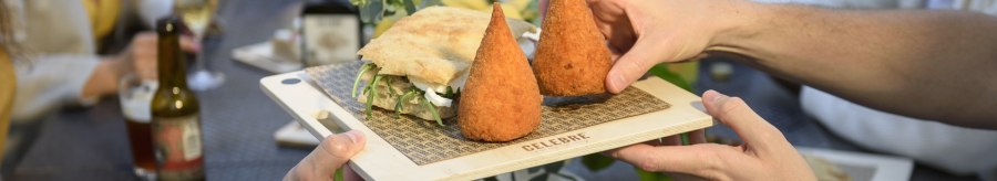 Two people pass a wooden board with arancini and a sandwich at a table with several guests., © CELEBRE