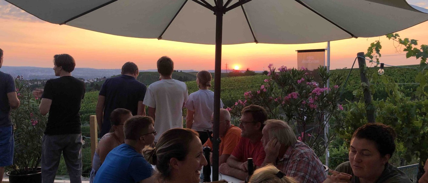 Menschen sitzen unter Sonnenschirmen in einem Weinberg und genie&szlig;en den Sonnenuntergang. Einige stehen und blicken in die Ferne., &copy; Weingut Diehl