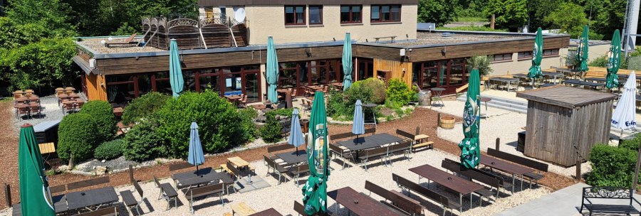 Outdoor area of Weitmann's Waldhaus with wooden tables, benches and parasols, surrounded by green trees and blue sky., &copy; Timo Weitmann