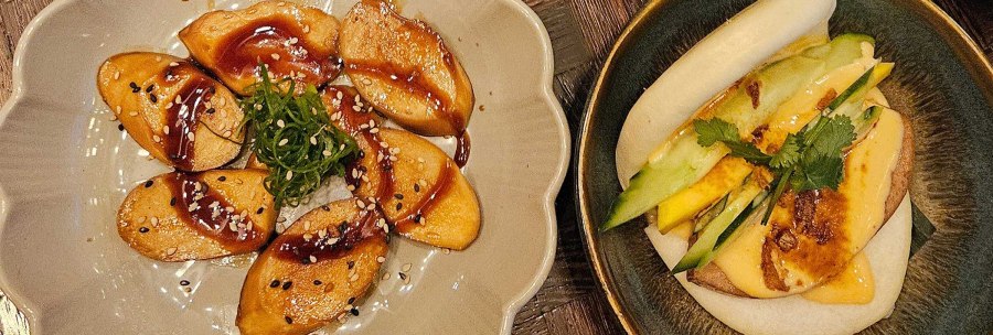 A plate of vegan dumplings in sauce, a bao bun with vegetables and a drink with ice cubes on a wooden table., © SMG A plate of vegan dumplings in sauce, a bao bun with vegetables and a drink with ice cubes on a wooden table., © SMG