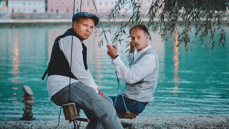Two men are sitting on swings on the riverbank, elegantly dressed. Water and buildings can be seen in the background., &copy; Rosenau Kultur e.V.