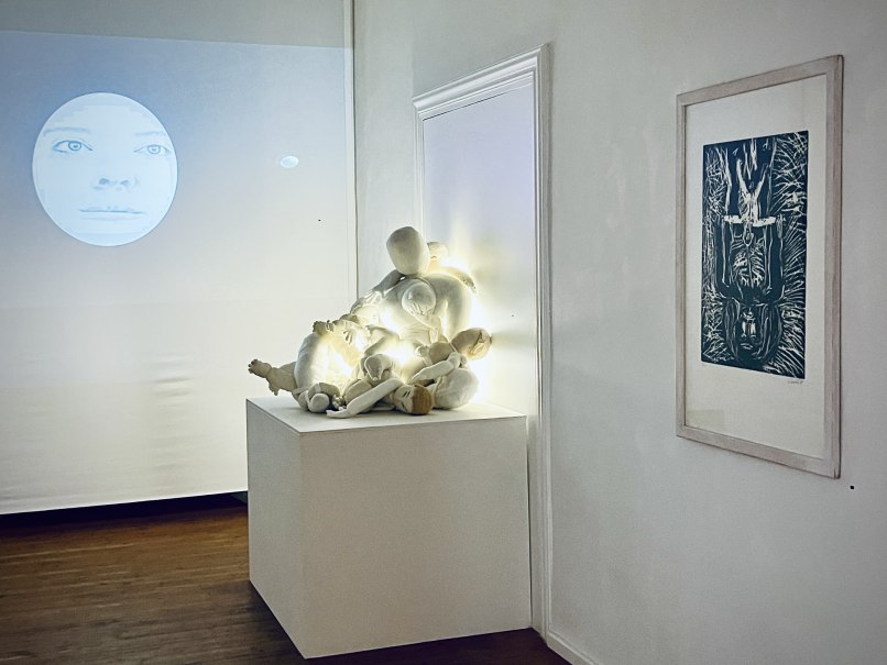 Eine Kunstinstallation zeigt eine Gesichtprojektion, beleuchtete Puppen auf einem Podest und ein gerahmtes Bild an der Wand., &copy; Min Bark