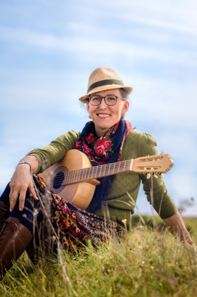 Eine Person mit Strohhut und Brille sitzt lächelnd auf einer Wiese, hält eine Gitarre und trägt bunte Kleidung., © Rosenau Kultur e.V. Eine Person mit Strohhut und Brille sitzt lächelnd auf einer Wiese, hält eine Gitarre und trägt bunte Kleidung., © Rosenau Kultur e.V.