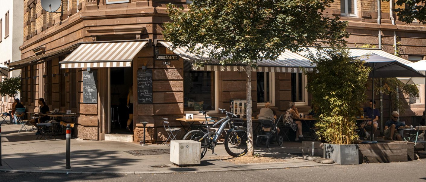 Ein gemütliches Straßencafé mit gestreiften Markisen, Tischen und Stühlen im Freien. Gäste sitzen im Schatten, Fahrräder sind abgestellt., © SMG, Sarah Schmid Ein gemütliches Straßencafé mit gestreiften Markisen, Tischen und Stühlen im Freien. Gäste sitzen im Schatten, Fahrräder sind abgestellt., © SMG, Sarah Schmid