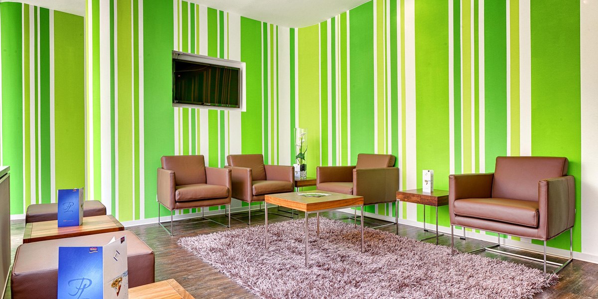 Moderne Lobby mit grünen Streifenwänden, braunen Sesseln und einem flauschigen Teppich. Ein Fernseher hängt an der Wand., © attimo Moderne Lobby mit grünen Streifenwänden, braunen Sesseln und einem flauschigen Teppich. Ein Fernseher hängt an der Wand., © attimo