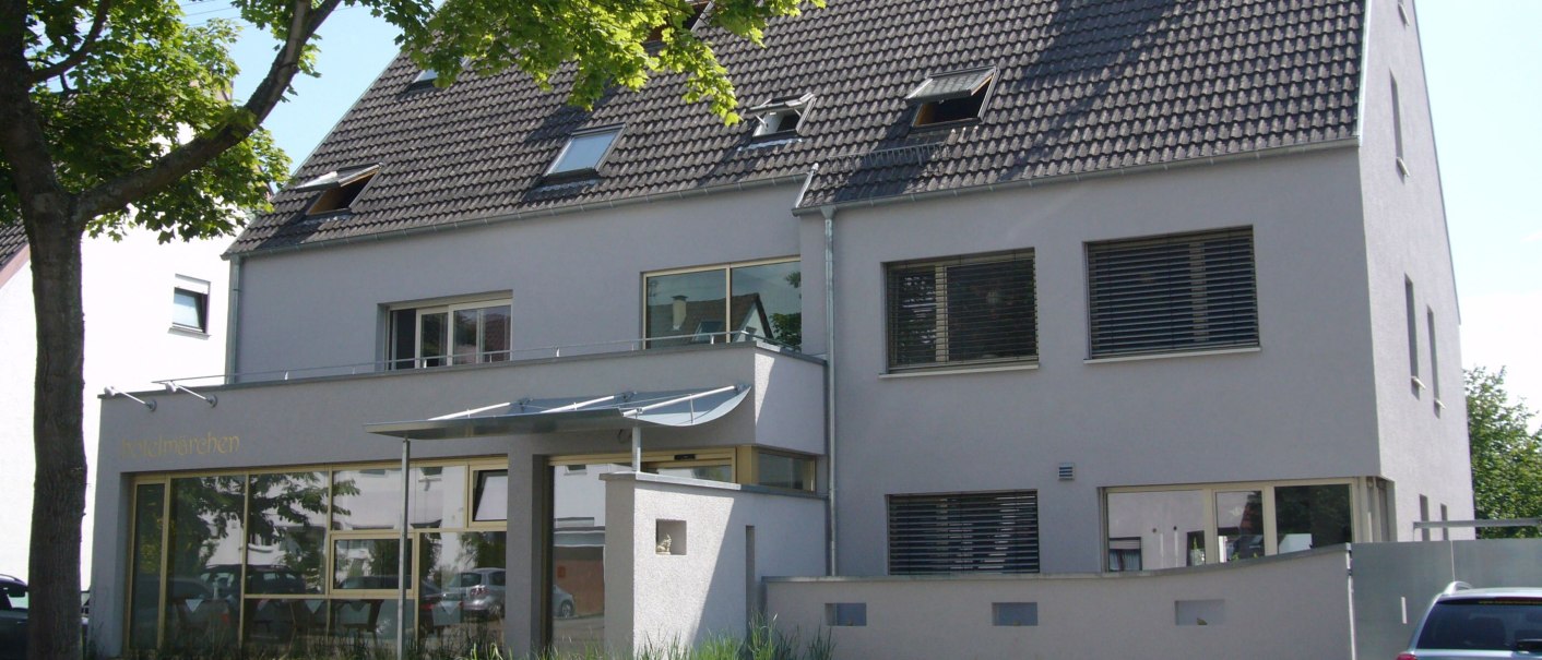 Modernes Gebäude mit grauem Dach, großen Fenstern und einem Baum im Vordergrund. Ein Auto parkt daneben., © hotelmärchen GmbH Modernes Gebäude mit grauem Dach, großen Fenstern und einem Baum im Vordergrund. Ein Auto parkt daneben., © hotelmärchen GmbH